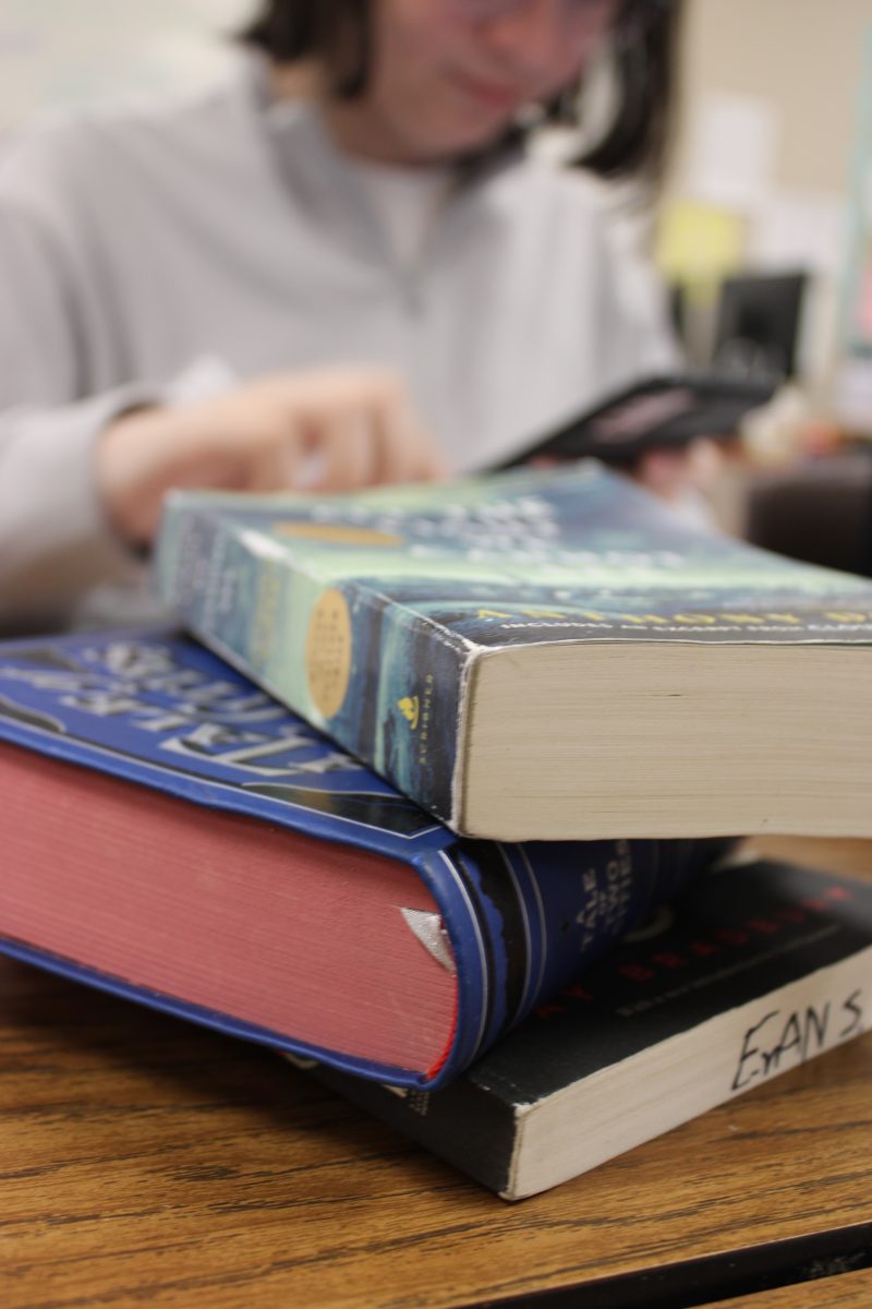 An AP student (Ryan Urbina) doing schoolwork in the background of Ms.Maimones classroom. 
Three novels sit in the foreground on a wooden school desk.